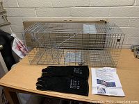 Photo of metal animal trap cage on table with black protective gloves and instruction sheet beside it