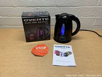 Black Ovente electric kettle with 1.7L capacity, original packaging, user manual, and a caution sign on a table against a concrete brick wall.