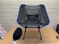 Portable fold-up travel chair set up on table with its black carrying bag beside it, chair has blue trim and mesh fabric seat with black metal legs.
