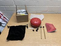 Photo of red steel tongue drum, two wooden mallets, five black tuning keys, black velvet bag, white cloth bag, instruction book and original box on wooden table against a light brick wall.