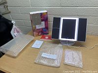 Two tri-fold light up vanity mirrors, one powered on showing LED illumination, complete with boxes, instruction manual, and protective bubble wrap on table.