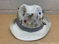 Front view of a white woven straw hat with a black and white checkered band decorated with various vintage pins including a peacock feather, rose, butterflies, and others.