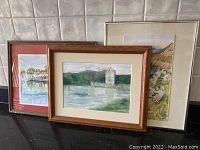 Three framed watercolour paintings: tower, bridge, and sheep scenes, shown together on a black surface against tiled wall