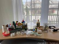 Photo of assorted craft supplies arranged on table including glue bottles, tape rolls, string spools, scissors, craft paints, glass jar and baskets