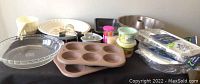 Kitchen table with baking pans and accessories arranged, showing the full assortment including metal and disposable muffin tins, pie and quiche plates, and other baking items.