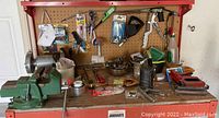 Wide view of workbench showing green bench vise on the left, assorted hardware and hand tools on pegboard and bench surface, and metal dual-wheel bench grinder at right.