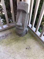 Front and right-side view of gray ceramic Moai-style statue on outdoor porch floor, showing its tall profile and detailed facial features.