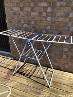 Full view of the foldable metal drying rack showing white metal rods and blue plastic connectors, placed on a wooden deck against a brick wall.