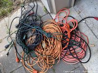 Assorted extension cords in black, orange, green and red on concrete surface, some coiled.