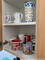 Photo showing assorted mugs in white cabinet, including ceramic travel mug and Bichon Frise dog mugs