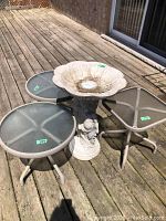 Photo of three small metal-framed tables with glass tops arranged outdoors next to a plastic birdbath with decorative base, showing condition and size relative to deck boards