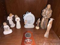 Photo showing religious figurines and decor on wooden shelf including angels, saint figurine, abstract figure, photo frame, and wooden plaque with holy water bowl