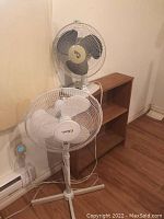 Photo showing two rotary fans - a white free-standing pedestal fan and a gray tabletop fan next to a small wooden shelving unit.