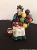 Front and side view of Royal Doulton The Balloon Seller figurine showing seated elderly woman with balloons and basket of flowers.
