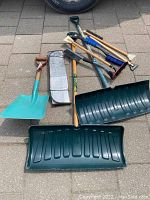 Full view of lot showing two large green snow push shovels, a smaller turquoise snow shovel with a D-grip wooden handle, several car snow and ice brushes with wooden handles, and a folded reflective sunshade for car windshield.