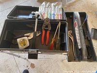 Black plastic toolbox with removable tray showing hammer, wire cutters, measuring tape, pliers, and two flathead screwdrivers