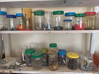 Multiple clear plastic jars with green, yellow, blue, and red lids filled with assorted screws, nails, washers, and hooks on shelves.