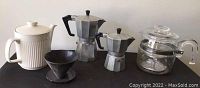 Full view of all coffee items in the lot on display on black surface against white wall showing Pyrex percolator, two aluminum Moka pots, Melitta filter, and stoneware carafe