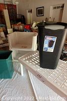 Photo showing white plastic shoe rack, black Rubbermaid garbage bin with lid, and green plastic storage tote on white surface.