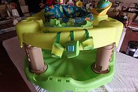 Front view of the baby play centre showing green plastic base, blue padded seat with animal print, and attached frog toy.