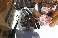 Photo showing two metal muffin tins, various cutlery and utensils in a plastic container, a wood salt and pepper mill, and patterned bowls arranged on a white tablecloth.