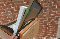 Photo showing assorted picture frames from side angle including black, white, and wooden frames leaning against a brick wall in a cardboard box.