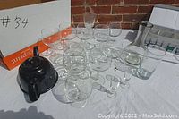 Overview of glassware items and black ceramic teapot arranged on white cloth, includes various stemmed glasses and a large glass carafe.