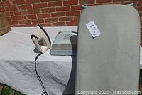 Black & Decker iron, ironing board, and boxed new ironing board cover displayed outdoors on a white cloth surface.