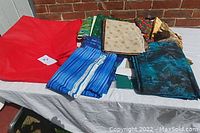 Wide view showing assortment of colorful sewing fabrics on a white table against a brick wall. Fabrics are varied in colors including red, blue, green, cream, and multicolored patterns.