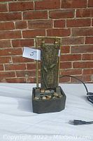 Front view of the electric water fountain showing decorative stones in a square base and vertical bronze-toned panel.