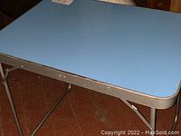Blue metal table top showing surface and edge detail with folding leg partially visible underneath.