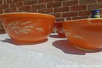 Three nested orange Pyrex mixing bowls with white interiors placed on a white cloth against a brick background. The largest has a white wheat pattern on the outside.
