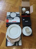 Lot overview showing boxed Alessi dinner plates, cups, and saucers on wooden floor.