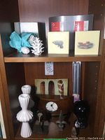 Photo of wooden Umbra mantle clock, two white ceramic candlesticks, white ceramic pine cone decor, two small square wall plaques, two silver metal photo frames, black round decorative item, and two arrow-shaped metal decor pieces on wooden shelf.