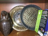 Two decorative metal plates and one carved pot on shelf, detailed brass-colored plate and black plate with Arabic calligraphy.