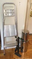 Full view showing ladder, two metal garbage cans, and manual air pump on hardwood floor indoors.