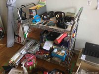 Full view of the Metro metal shelving cart filled with various items showing wire shelves and glass middle shelf with wheels on floor.