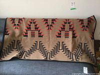 Front view of the wool blanket draped over a couch, showing geometric patterns in red and black on tan background.