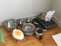 Full set of cookware and bakeware showing three stainless steel pots with glass lids, a stainless steel frypan and small pot, metal strainer, glass baking dish, Brita water jug, and metal baking trays.