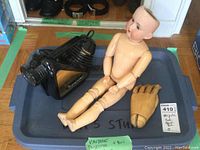 Wide view showing the black Magnajector projector, vintage nude doll missing hair with jointed limbs, and wooden hand all placed in a plastic bin.