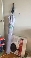 Patio sun umbrella wrapped in gray cover with crank handle leaning against wall next to boxed umbrella base