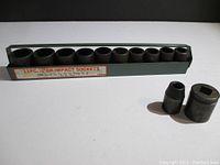 11 impact sockets held in a metal tray with two sockets placed separately outside the tray on a black surface, showing front and side angles of the sockets and tray with label detailing sizes.