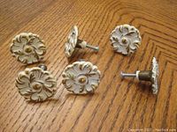 Five vintage white and gold floral design cabinet knobs arranged on wood surface showing front and side views, screws attached to knobs visible.