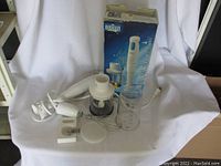 White Braun Multiquick hand blender with power cable, blending wand, whisk attachment, chopping bowl with lid, measuring cup, and original retail box placed on white cloth background.