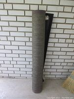 Rolled up outdoor rug leaning against a white brick wall, showing texture and color as dark brown.