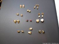 View of 12 pairs of various vintage clip-on earrings spread out on a black surface showing different gold-tone, faux pearl, and rhinestone designs.