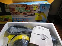 Opened box showing yellow and black steam cleaner unit with hose and funnel, user manual included beside the unit.