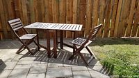 Full view of Ikea Applaro folding outdoor table and two matching folding chairs on patio blocks in sunlight.