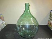 Front view of large green glass bottle with rounded body and narrow neck on table against wall.