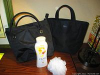 Two black tote bags standing on a wooden surface, one labeled Olay with handles and metal rings, the other BCBG with short handles, with the Olay body wash bottle and white bath pouf placed in front.